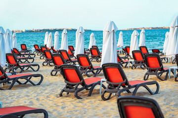 Rows of red lounge chairs line the sandy shore of a beach, each positioned under a white beach...