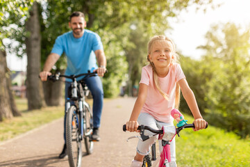 Obraz premium A young girl is smiling as she rides a pink bicycle on a paved path in a park. Her father is behind her, also riding a bicycle. Both are enjoying a sunny day outdoors.