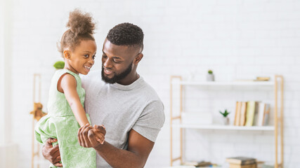 Obraz premium African American father smiles and holds his daughter in his arms, both looking at each other. The background features a bookshelf with books and decor in a white home interior.