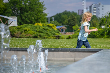 Young Girl Running Past a Fountain in a City Park on a Sunny Day. Pure Happiness concept: Focusing on the pure and unfiltered happiness of children at play.