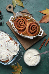 Baking dishes of tasty cinnamon rolls with cream and autumn leaves on green background