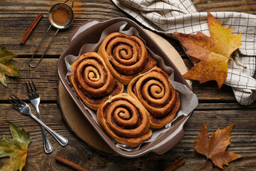 Baking dish of tasty cinnamon rolls and autumn leaves on wooden background