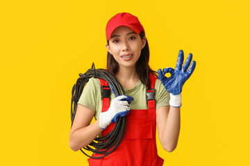 Young female electrician with cables showing OK gesture on yellow background
