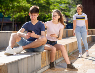 Interested teen boy sitting with phone on summer city street with girlfriend trying to get his attention. Concept of phubbing behavior..