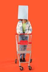 Beautiful young stylish woman with trolley and shopping bag on orange background