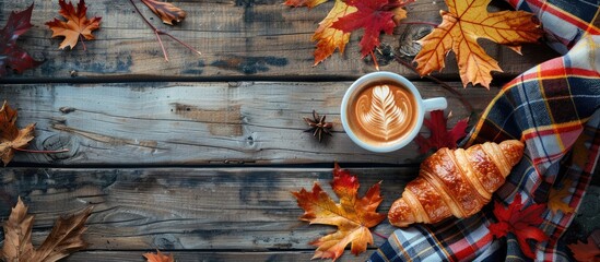 Autumnal concept portrayed in an image featuring a cozy setup with a cup of cappuccino, a croissant, plaid, and maple leaves spread out on a wooden surface in a flat lay composition with copy space