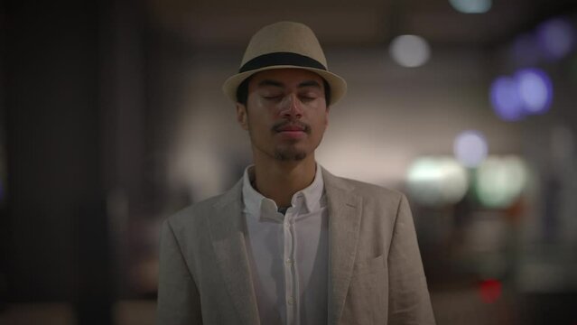 An elegant gentleman with a hat on, peacefully shutting his eyes, set against a blurred background