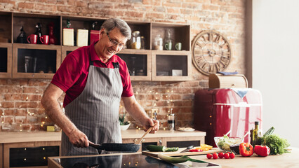 A smiling senior man wearing an apron cooks in a modern kitchen with brick walls. He is stirring...