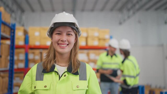 Beautiful young warehouse woman worker stand in front of her co-worker men discuss together near product shelves and look at camera with smiling.