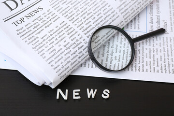 Newspapers with magnifying glass and word NEWS made of letters on black wooden background
