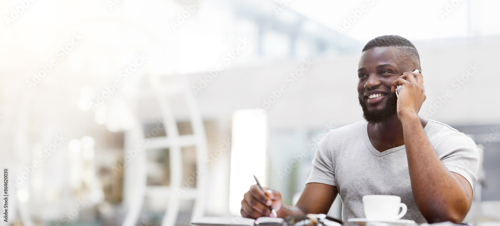 Poster a man sits at a table in a cafe, talking on his phone and taking notes. he is smiling and looks happ