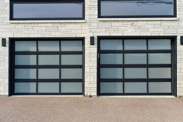 Two large car garage doors on a contemporary style house. The black metal double doors have glass panels and two windows above the entrance of the grey stone building. The driveway surface is concrete