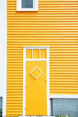 A bright yellow house with white trim. The exterior wall is made of narrow pine clapboard siding. There's a colorful single shutter door with a diamond frame and a transom window over the entrance.