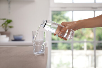 Woman pouring water from bottle into glass on table in kitchen, closeup