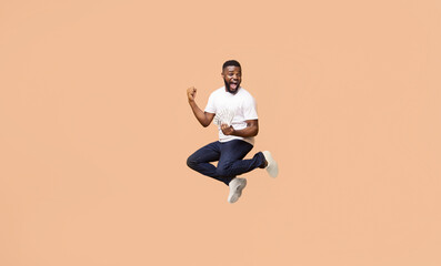Happy, young black man jumping in air. He is wearing a white t-shirt, blue jeans, and white sneakers. He is holding a wad of cash in his left hand and his right arm is raised in the air, copy space
