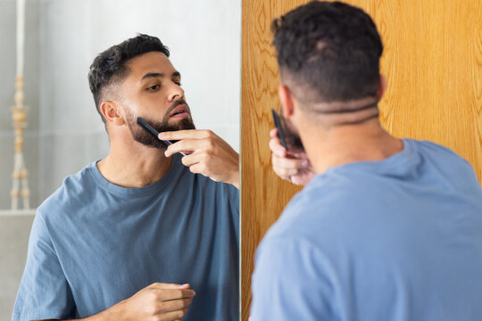 Grooming beard, man using comb and mirror for precise styling at home