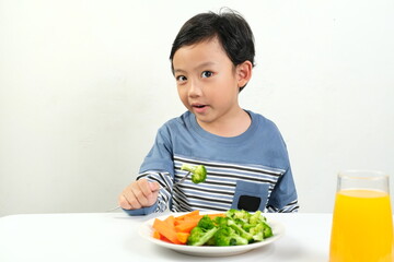 A boy in a blue shirt enjoys a healthy meal with broccoli, carrots, and juice. His curious expression emphasizes the importance of nutritious eating.Ideal for health promotions and educational content