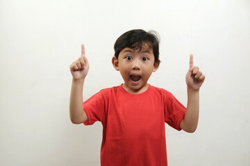 A surprised young boy in a red shirt points upward with both hands, his eyes wide open and mouth...