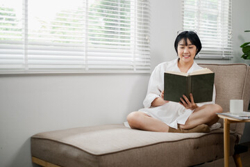 A woman enjoys a peaceful moment reading a book on a cozy sofa in a well-lit, modern living room with large windows and white blinds.