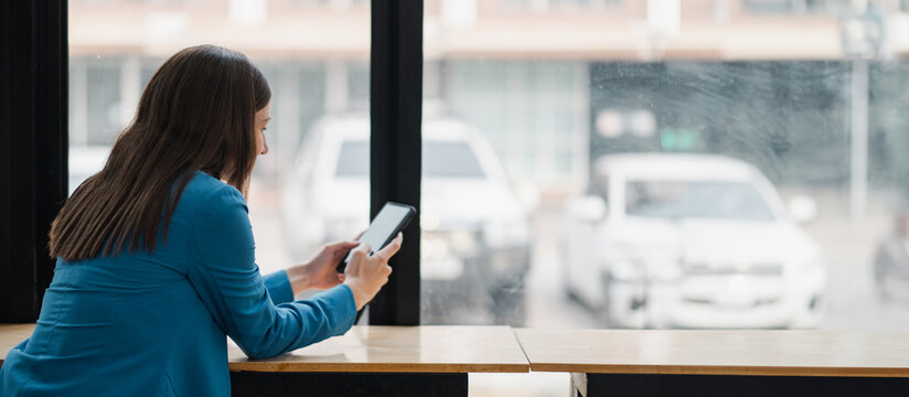 Woman in blue jacket using a tablet by a window in a modern cafe, with cars visible outside. Casual business environment.