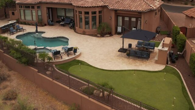 An aerial 4k video of a Deseret landscaped backyard in Arizona featuring a travertine tiled pool deck with a spa, outdoor fireplace, outdoor kitchen and putting green.
