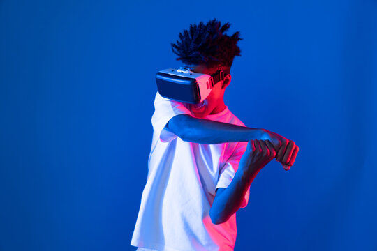 Young African American player wearing VR playing sword or baseball hologram isolated blue pink neon lighting wall connecting digital futuristic technology virtual reality metaverse world. Contrivance.