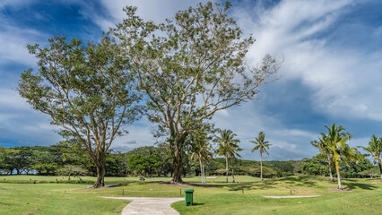 Footpaths meander between golf courses. The lawns have trimmed green grass, palm trees, deciduous. In the distance there are golf carts, silhouettes of people playing. Blue sky, clouds. Malaysia.