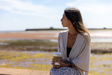 Young woman holds hot tea sitting in front of ocean on windy day 