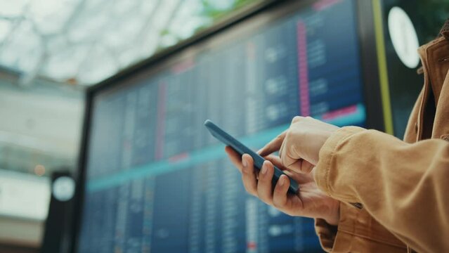 Hands of traveler using smartphone checking his flight status and self check-in nearby arrivals board, travel and tourist in airport and transportation