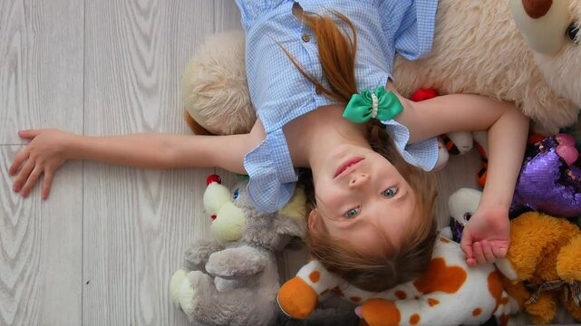 A charming little girl with pigtails is lying on the floor among scattered soft toys, looking into the frame with big blue expressive eyes, smiling. A child's free play.