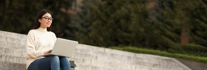 Asian young woman sits on concrete stairs outside, working on a laptop. She is wearing a white sweater and jeans, and has long, dark hair, copy space