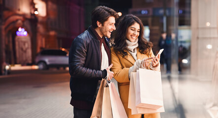 Young couple with smartphone near shopping mall, standing near shop window in the evening