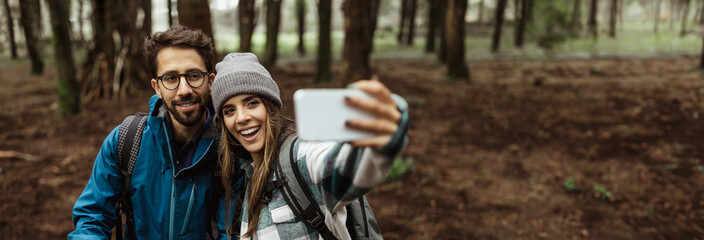 A young couple, dressed in casual outdoor clothing, stands in a forest and takes a selfie with a smartphone. They are smiling and seem to be enjoying their time together, copy space