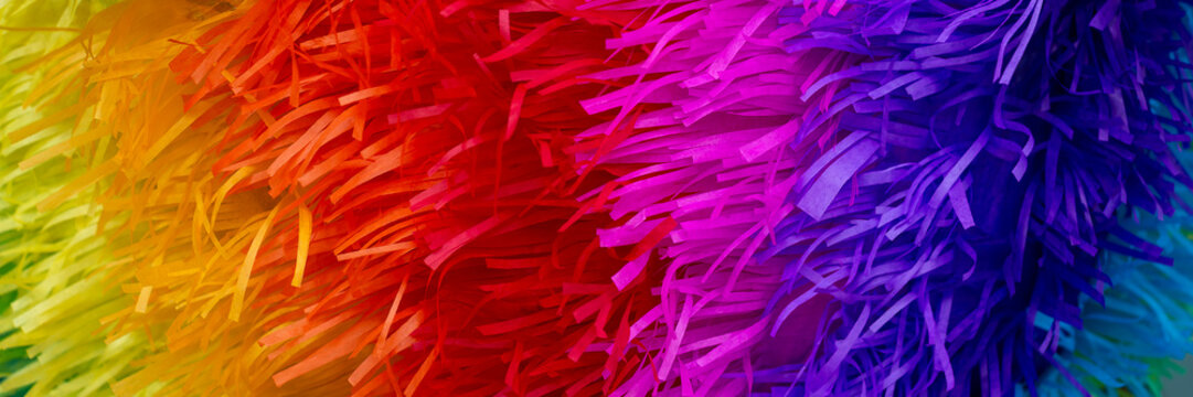 Bright rainbow colored paper, abstract background texture of a pinata. Close-up, multicolored, fringed paper in yellow, orange, red, pink and purple. Banner sized, design element with copy space.