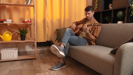 Obraz premium Adult man guitarist sitting in the room playing acoustic guitar, composing melody, fingering chords. Hobby and leisure time concept.