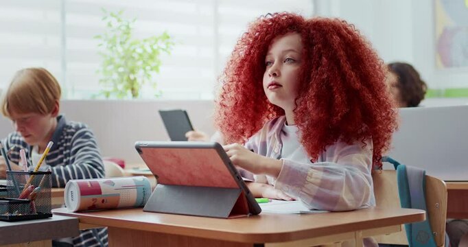 Young Caucasian girl with long red hair studying in classroom. Using tablet device. Modern technology. Writing down something on touch screen with special pen. Listening to teacher.