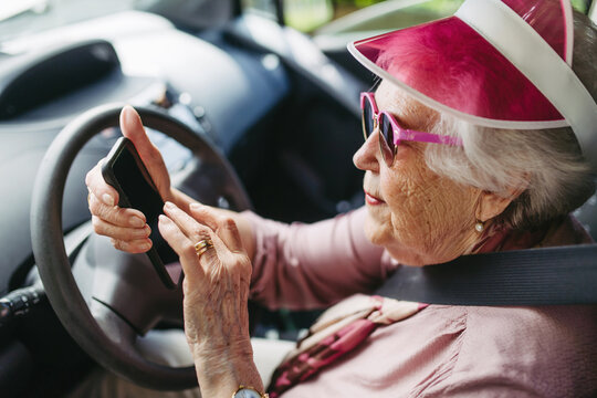 Happy senior woman driving car alone, holding smartphone and scrolling. Safe driving for elderly adults, older driver safety.