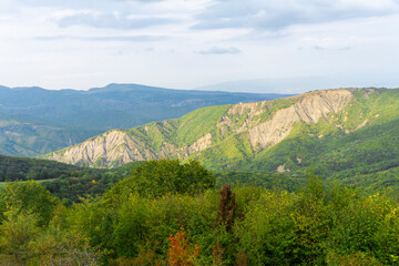 Naklejka premium The rock, partially overgrown with bushes, is illuminated by sunlight. Green trees in the foreground. Mountains and sky in the background