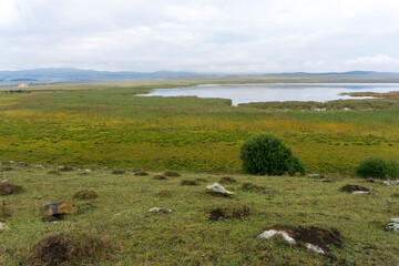 Bareti lake surrounded by fields of faded and green grass. Rocks and bushes in foreground