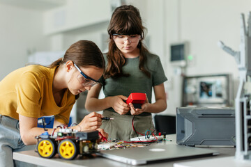 Fototapeta premium Two girl classmates working togeter on circuit board, building robotic car in after-school robotics club. Children learning robotics in Elementary school.