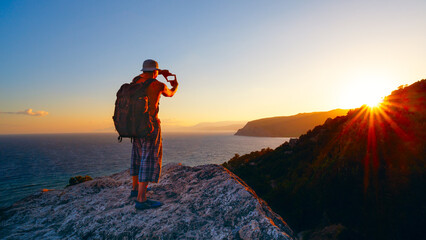 World Photography Day, man takes a photo of a sunset with a backpack. The scene is peaceful and serene: a man stands on a rocky hill overlooking the ocean. The sun sets on the mountains