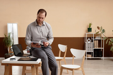 Minimal full length portrait of adult businessman reading document leaning on desk in modern office workspace copy space