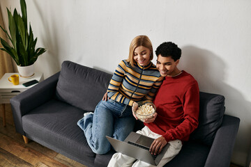 A happy multicultural couple cuddles on a sofa in a modern apartment, enjoying a movie night together with popcorn and a laptop.