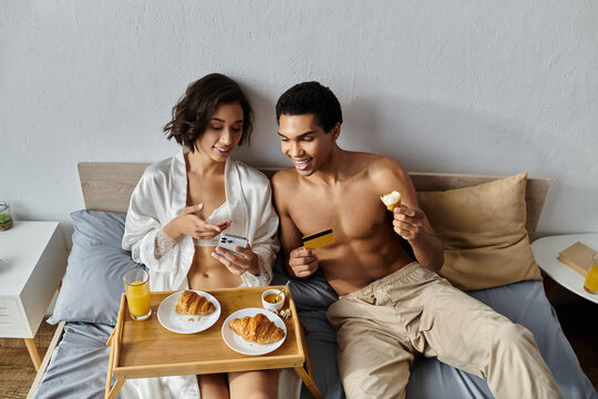 A young couple enjoys a leisurely breakfast in bed, with the woman checking her phone and the man taking a bite of pastry. - Powered by Adobe