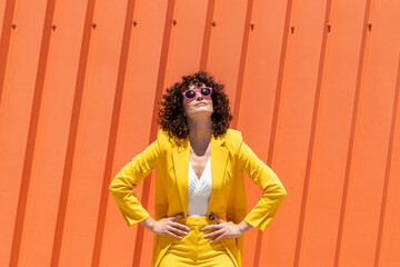 Woman with arms akimbo wearing sunglasses and yellow blazer in front of orange wall on sunny day