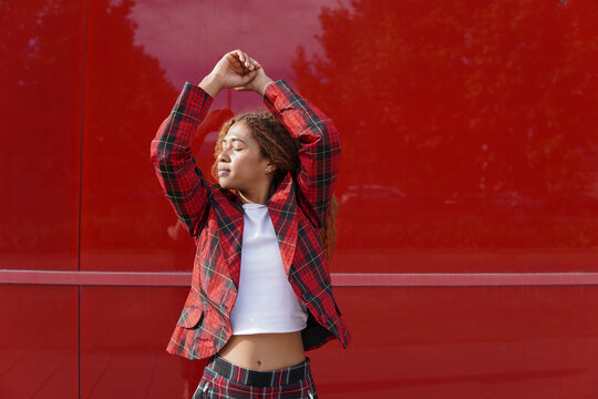 Young woman wearing checked blazer and standing with arms raised in front of red wall