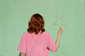 Redhead woman wearing pink shirt holding star shape in front of mint green wall on sunny day