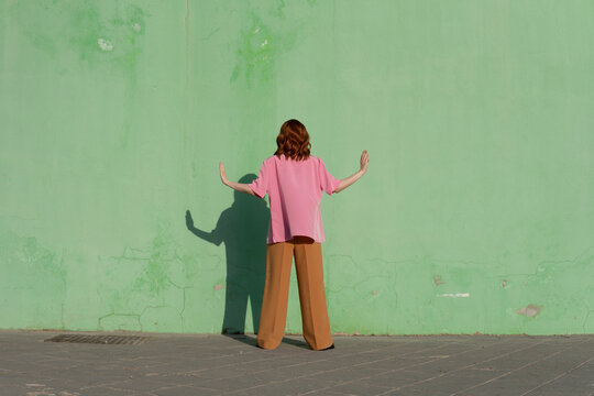 Redhead woman standing with arms outstretched in front of mint green wall on sunny day