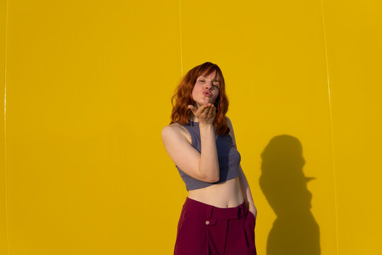 Beautiful redhead woman blowing kiss standing in front of yellow wall