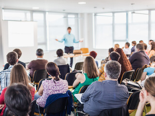 Business and entrepreneurship symposium. Speaker giving a talk at business meeting. Audience in conference hall. Rear view of unrecognized participant in audience.